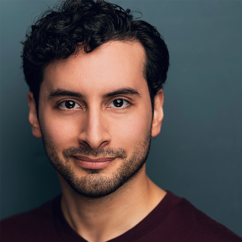 A headshot of a performer with dark curly hair and light stubble looking directly at the camera with a gentle smile. He is wearing a dark maroon shirt against a plain, muted blue-grey background.