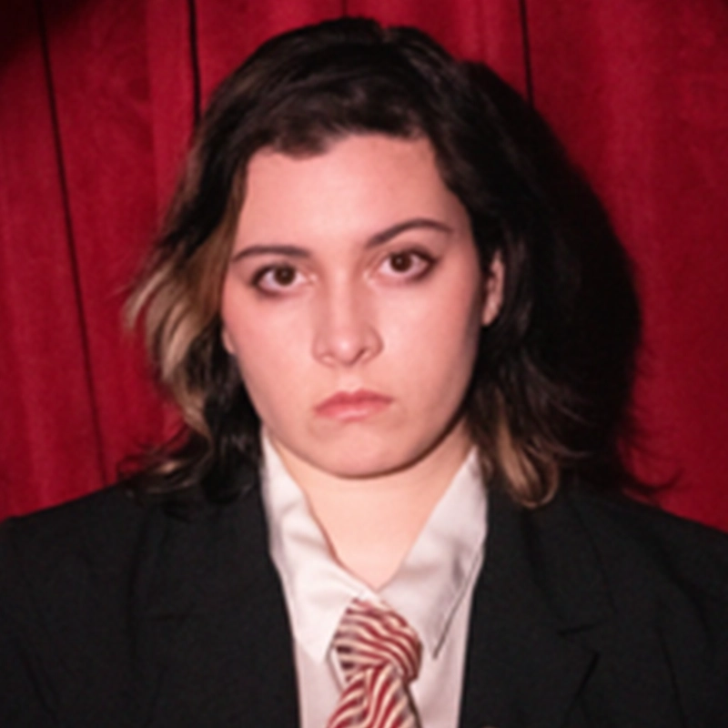 Sofia R. C. Melendez, with wavy brown hair and a streak of blonde, stands in front of a red curtain wearing a black blazer, white shirt, and striped tie. Their expression is serious as they look directly at the camera.