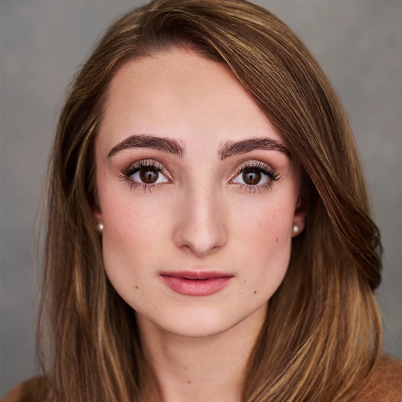 Headshot of Sophia Manicone with long, light brown hair looking directly at the camera. She has a neutral expression and is wearing small stud earrings.