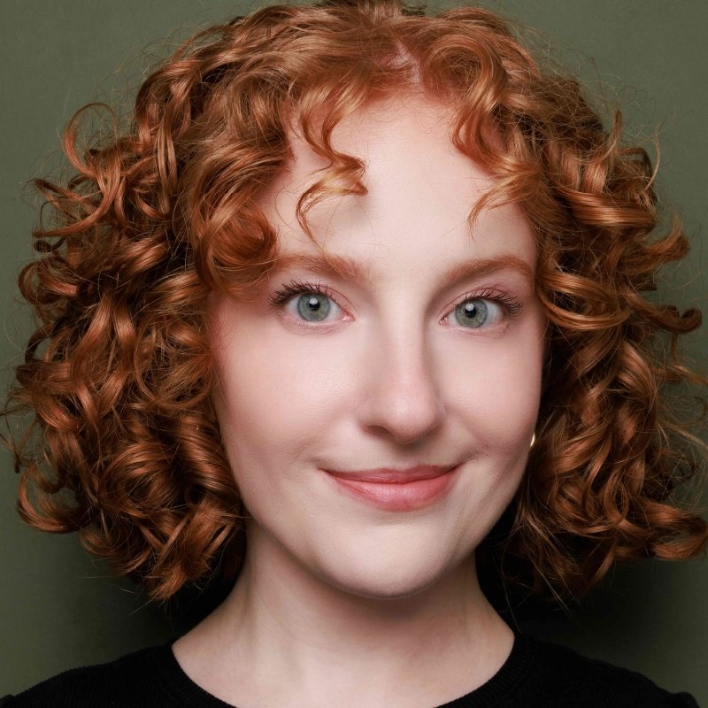 Headshot of Sophia Manicone with shoulder-length curly red hair looking directly at the camera with a gentle smile. They have light skin and light-colored eyes, set against a plain olive-green background.