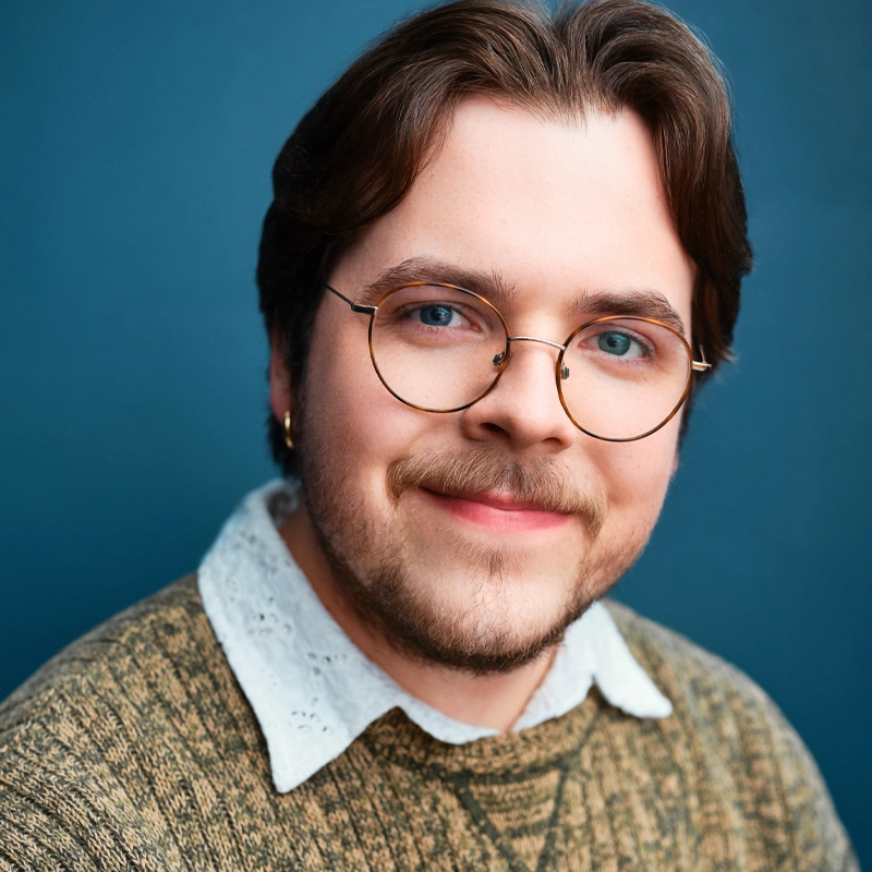 Tristan Tierney, with light skin, brown hair, and a trimmed beard, smiles softly. They wear round glasses, a green knit sweater over a white lace-collared shirt, and a small earring against a solid blue background.