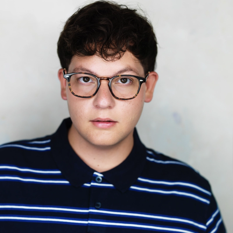Young person with short curly brown hair and glasses, wearing a navy blue collared shirt with white horizontal stripes, stands against a light, neutral background, looking directly at the camera with a neutral expression.