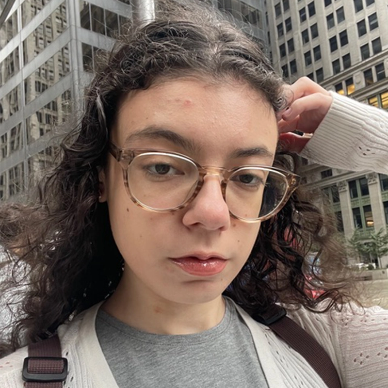 Sofia R. C. Melendez, with curly brown hair, clear glasses, and a neutral expression, stands outdoors in a city of tall office buildings. She wears a light sweater over a gray top and touches her head with one hand.