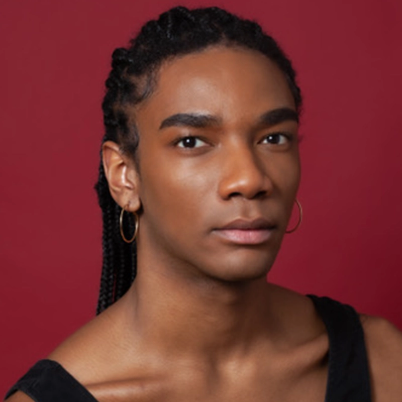 Sofia R. C. Melendez, with medium-dark skin, long braided hair, and gold hoop earrings, looks directly at the camera. She wears a sleeveless black top and has a neutral expression against a solid deep red background.