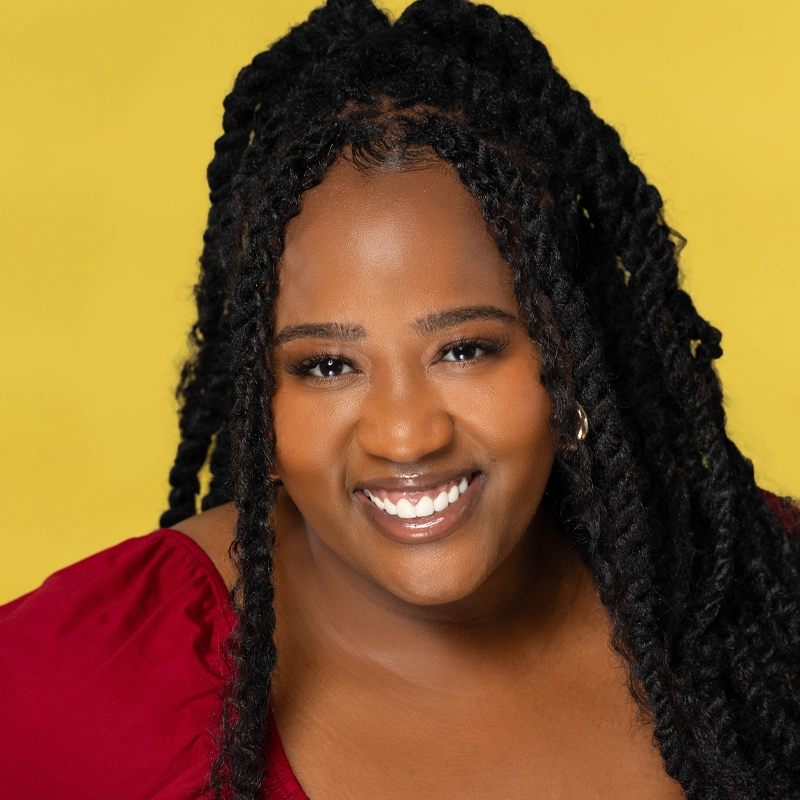 Aneesa Folds, with long, textured black hair styled in twists, smiles warmly at the camera. She wears a deep red top and small hoop earrings against a solid, vibrant yellow background, creating a cheerful and lively atmosphere.