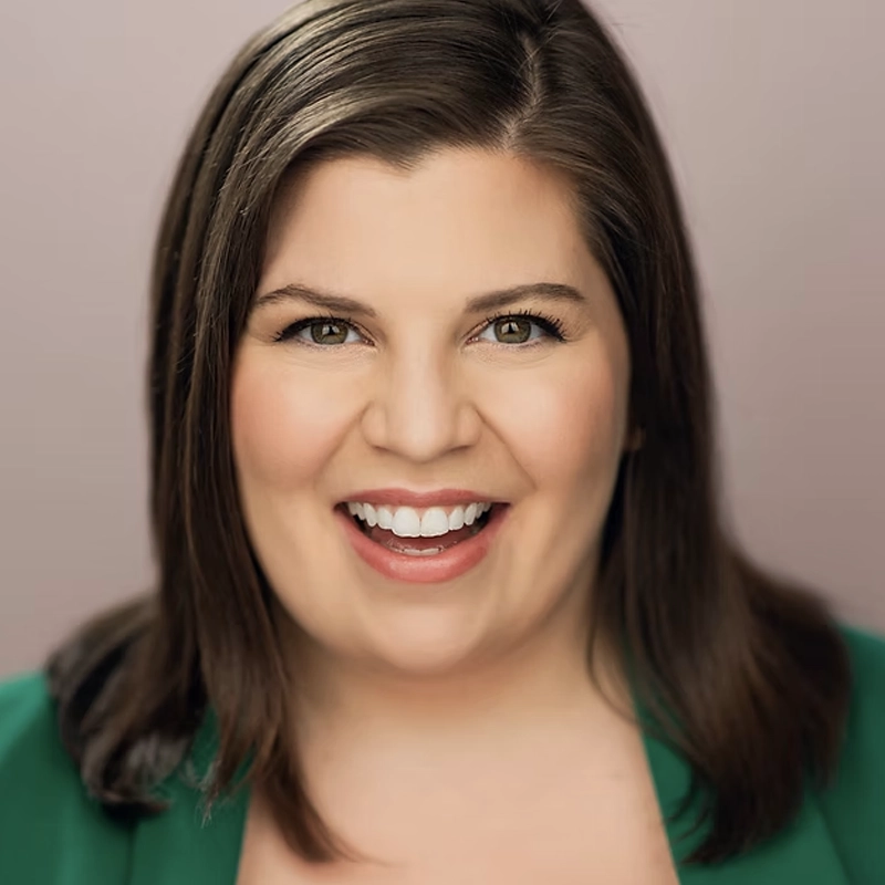 Katelyn Lauria, with straight brown hair and fair skin, smiles warmly at the camera. She wears a green blazer over a light top, while the softly blurred neutral background draws focus to her expressive face and friendly demeanor.