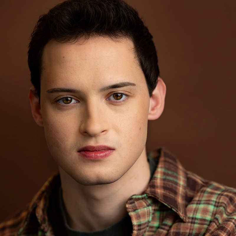 Noi Nuria Maeshige, a young person with fair skin, short dark hair, and brown eyes, looks directly at the camera. They wear a plaid shirt over a dark top against a warm brown background, creating a soft and neutral portrait setting.