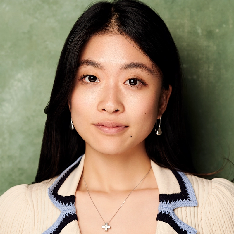 A young woman, Noi Nuria Maeshige, with long black hair, wearing a cream cardigan with dark trim, pearl drop earrings, and a silver cross necklace, poses against a textured green background. She has a natural expression and a beauty mark near her mouth.