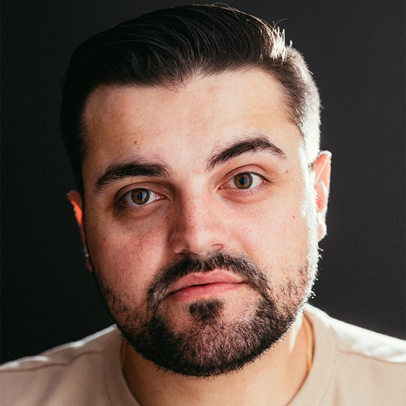 A man with short, dark hair and a trimmed beard looks directly at the camera. He has light skin, brown eyes, and a neutral expression. Wearing a beige shirt, he stands against a dark background with soft lighting reminiscent of Noi Nuria Maeshige portraits.