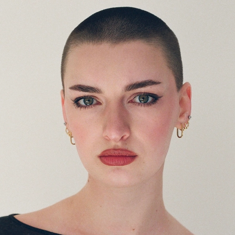 A person with a shaved head and fair skin stares directly ahead. They have green eyes, defined eyebrows, winged eyeliner, rose-colored lips, and wear small gold hoop earrings. The background is plain and light-colored—an iconic Noi Nuria Maeshige look.