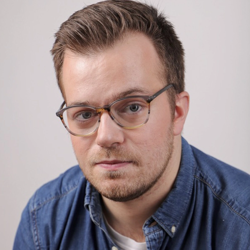 A young man with short brown hair, beard, and mustache wears rectangular eyeglasses and a blue denim shirt over a white t-shirt. Derek Speedy looks directly at the camera with a neutral expression against a plain, light-colored background.