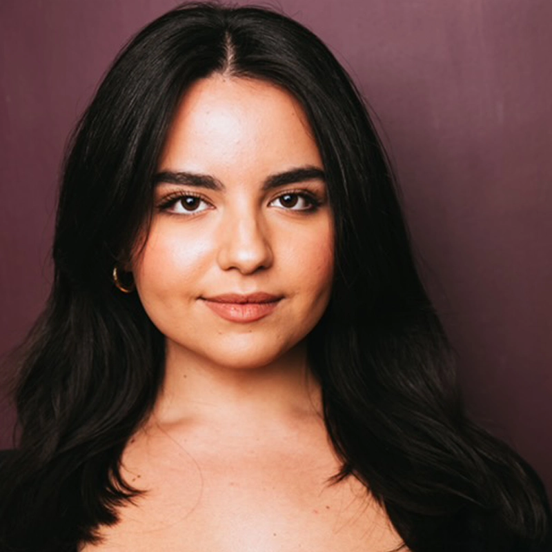 A young woman with long, dark hair and medium skin tone is facing the camera, smiling softly. She wears subtle makeup, gold hoop earrings, and a black top. The background is a solid, deep purple color. The lighting is even and flattering, capturing Isabella Walther-Meade’s poised presence beautifully.
