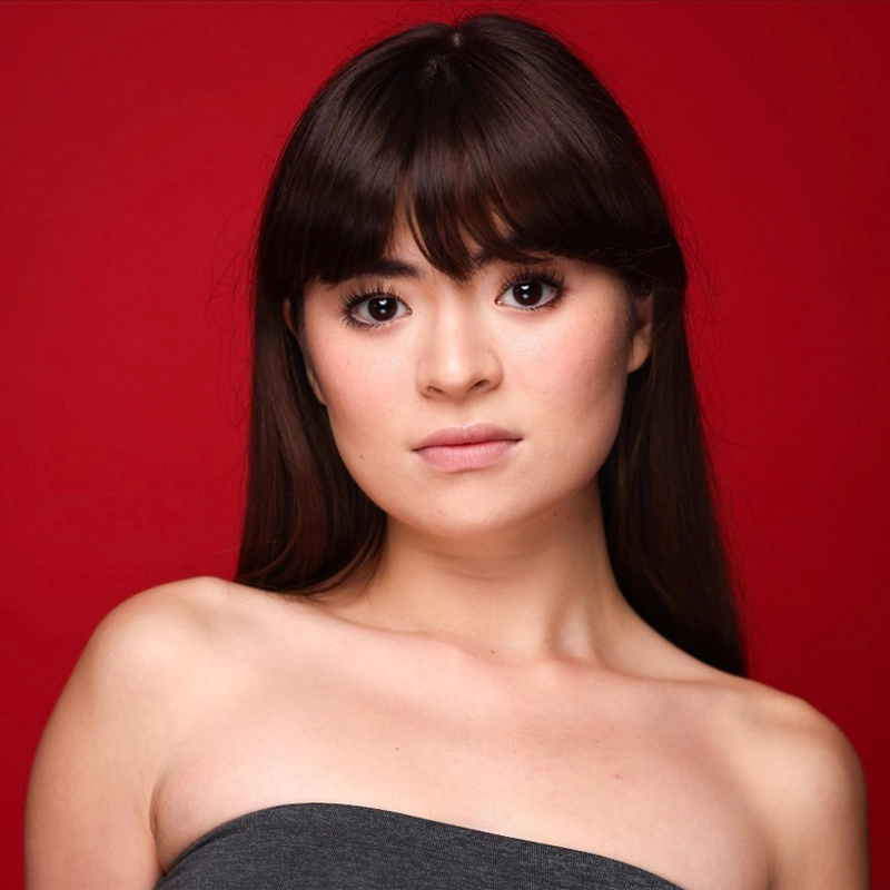 A young woman with long dark hair and bangs poses against a solid red background. She wears a gray, strapless top and looks directly at the camera with a neutral expression. Her skin is fair, and she has subtle makeup, reminiscent of Anna Zavelson’s understated elegance.