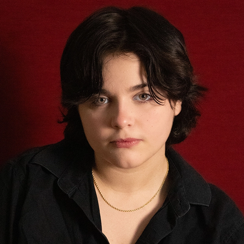 A young person with short dark hair and light skin, wearing a black shirt and a thin gold chain necklace, looks directly at the camera with a neutral expression. The background is a solid deep red color.