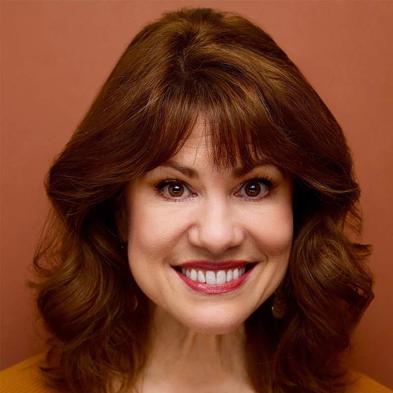 Michelle Jennings, a woman with wavy, reddish-brown hair and bangs, smiles warmly at the camera. She wears natural makeup and a brown top against a solid, warm brown background that complements her hair and outfit.