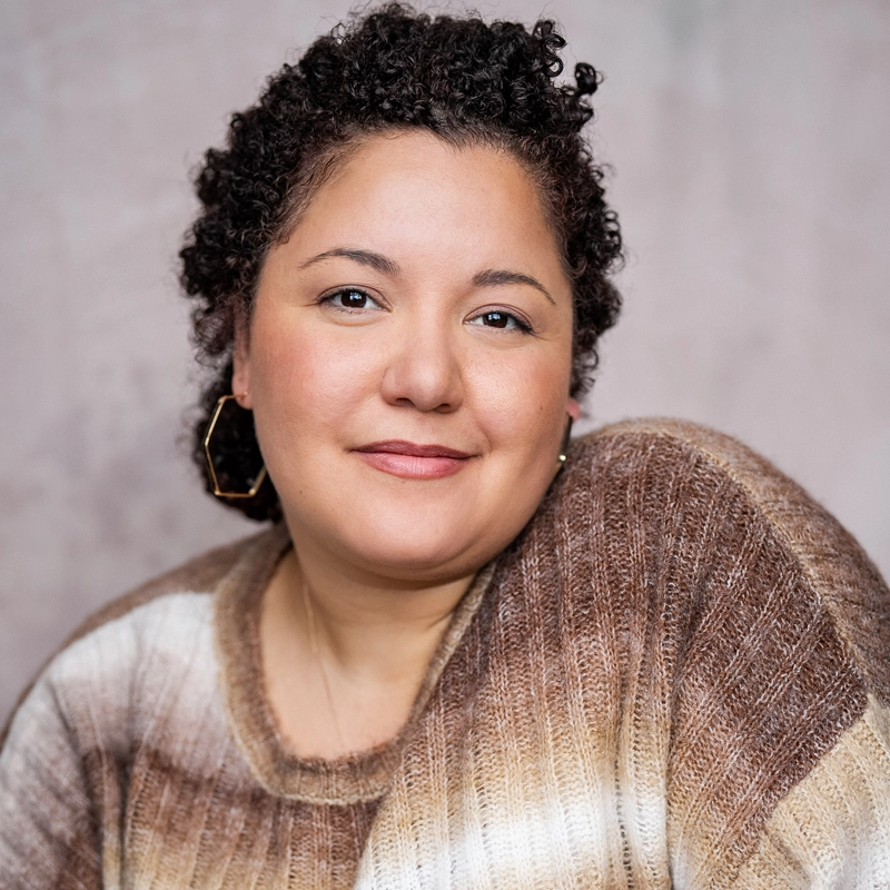 LaRaisha Dionne, a woman with curly dark hair and medium skin tone, gazes softly at the camera. She wears a striped brown and cream sweater and large geometric earrings, her gentle expression framed by a neutral blurred background.