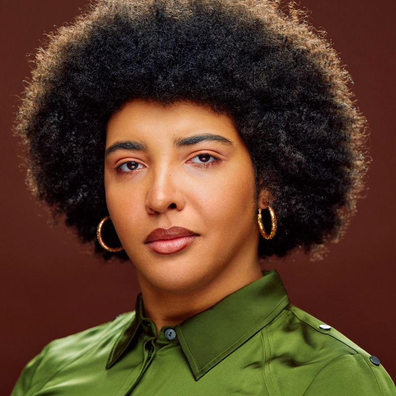 A person with a medium-dark complexion and a voluminous curly afro hairstyle is shown against a brown background. They wear gold hoop earrings and a shiny olive-green button-up shirt, and their expression is calm and confident.