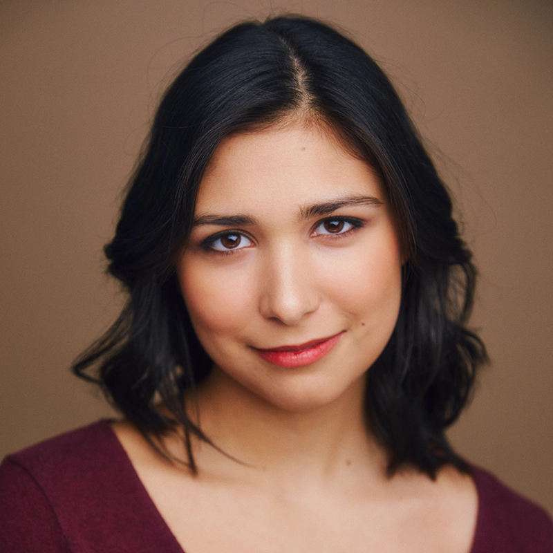 Isabella Orosco, a young woman with medium-length dark hair and warm-toned skin, smiles softly. She wears a burgundy top and natural makeup with subtle red lipstick. The smooth, neutral brown background and soft lighting highlight her relaxed expression.