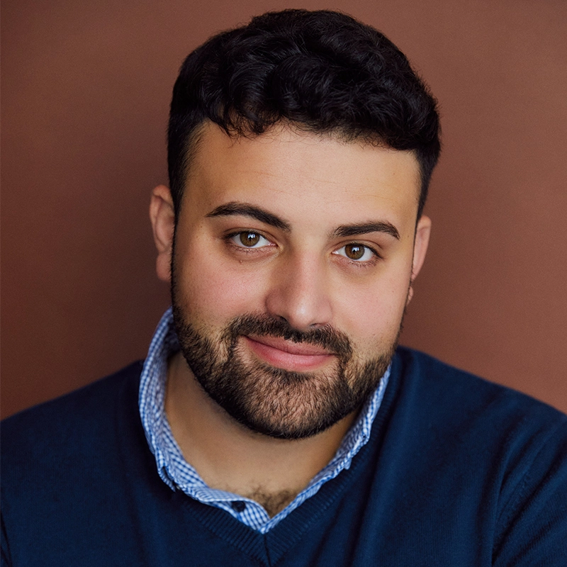 A person with short, dark curly hair and a trimmed beard smiles gently. They wear a navy sweater over a blue checkered shirt. The background is a solid, warm brown color. The lighting is soft, highlighting their friendly expression.