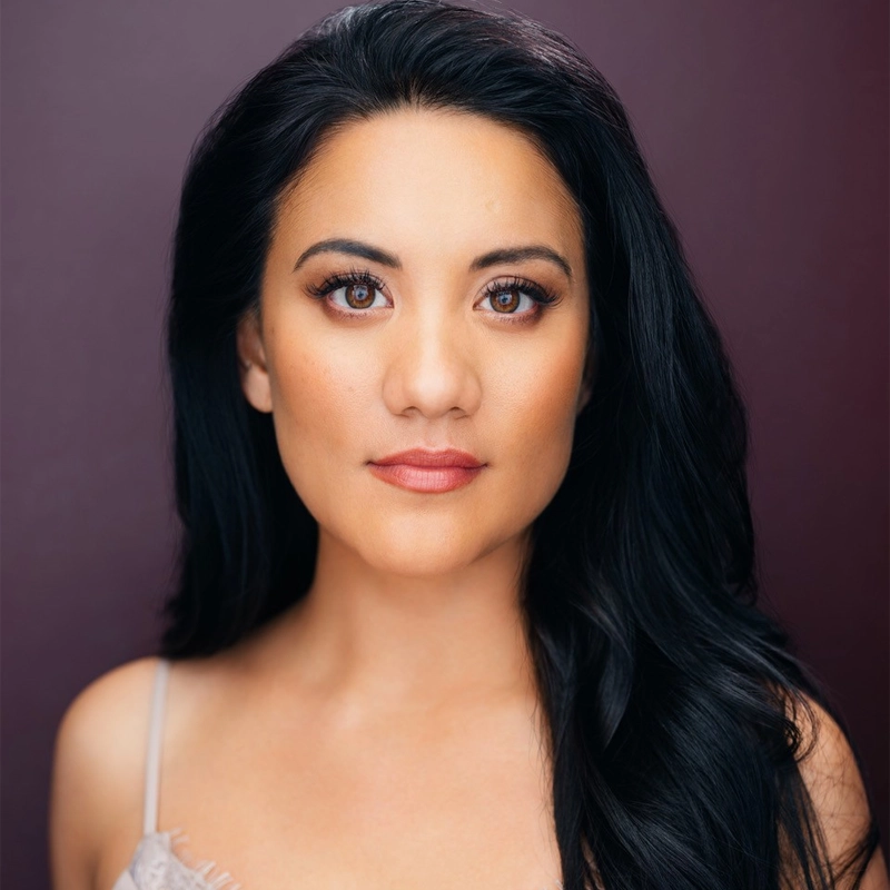 A woman with long black hair and smooth skin stares confidently at the camera, wearing natural makeup and a beige thin-strapped top, against a deep plum background. Her calm, self-assured expression is reminiscent of Diane Phelan’s poised presence.
