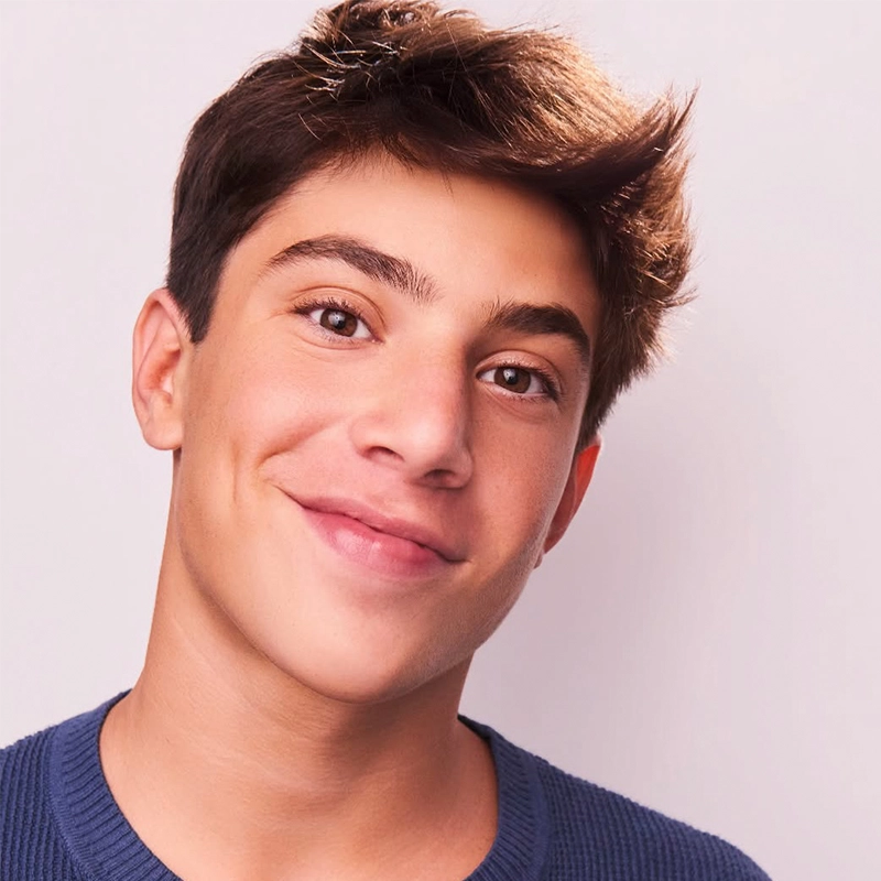 A teenage boy with short, dark brown hair smiles at the camera. He wears a blue textured crewneck sweater and stands against a light pink background. His head is slightly tilted, and his expression is warm and friendly—capturing the approachable charm often associated with Remi Tuckman.