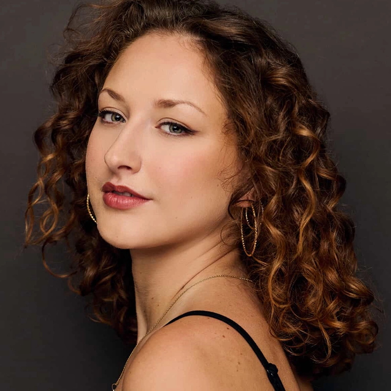 Valentina Perdido, with light skin and curly, shoulder-length brown hair, looks over her shoulder. She wears gold hoop earrings, a necklace, subtle makeup, and a black strap on her shoulder against a dark gray background.