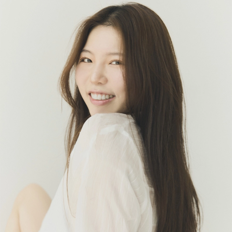 A young woman with long, straight dark hair sits sideways, turning her head to smile at the camera. Joo Won Shin wears a sheer, white top and is set against a plain light background, creating a soft and natural portrait.