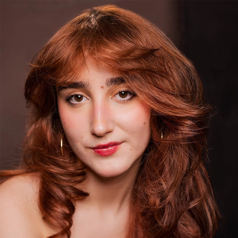 Valentina Perdido, a young woman with long, wavy auburn hair and fair skin, looks at the camera with a gentle expression. She wears natural makeup, pink lipstick, and gold hoop earrings against a softly blurred dark background.