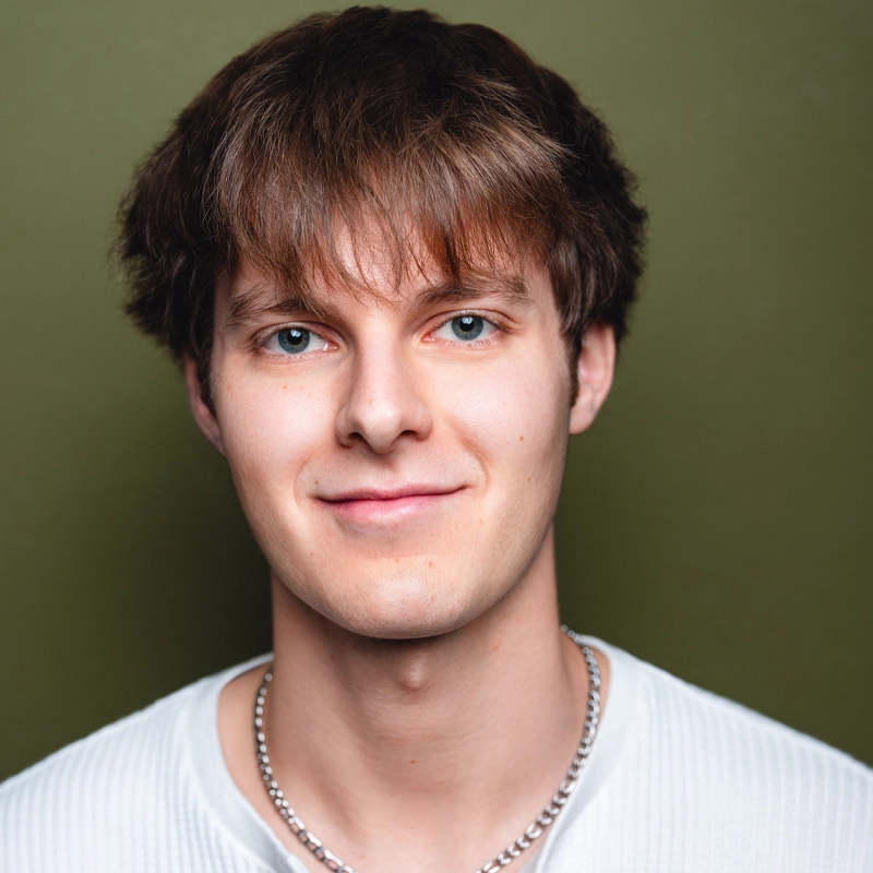 A young man with fair skin and straight brown hair smiles softly at the camera. He wears a white ribbed shirt and a silver chain necklace. The background is a solid muted green color, giving the portrait of Ryan Bronston a serene and modern feel. The lighting is bright and even.