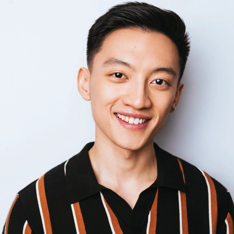 A young man with short black hair smiles at the camera. He is wearing a black collared shirt with vertical brown and white stripes. The background is plain and light-colored, giving the portrait a clean and bright look.