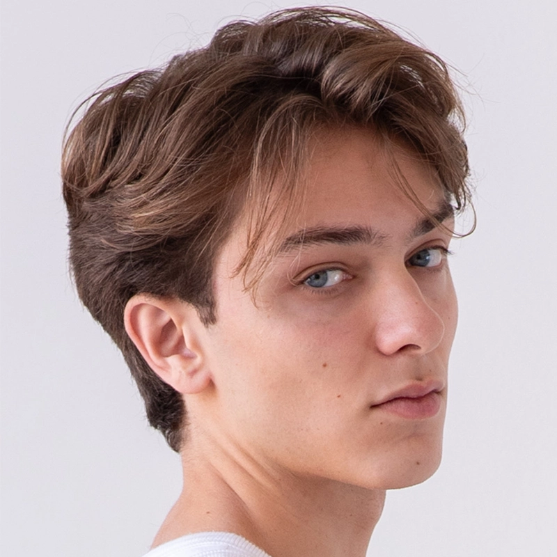 Young person with light skin and tousled brown hair gazes over their shoulder toward the camera, wearing a white shirt. The plain, light background draws focus to Noah Ong Bamola’s neutral facial expression.