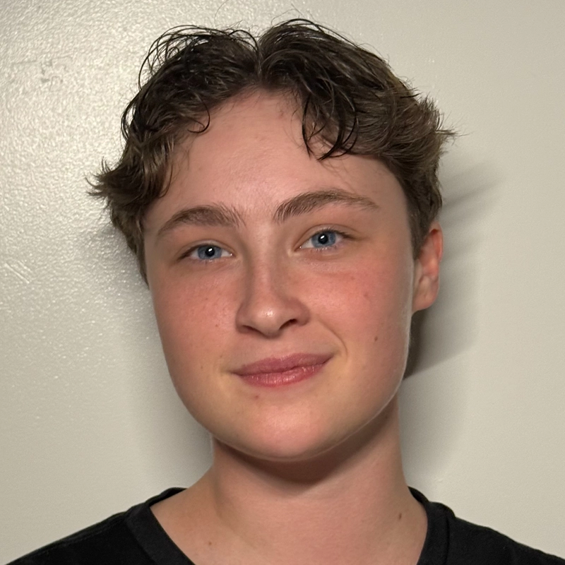 A young person with short, wavy brown hair and blue eyes smiles gently in front of a plain off-white wall. They wear a black shirt and have fair skin, with soft lighting creating subtle shadows behind them for a calm atmosphere.