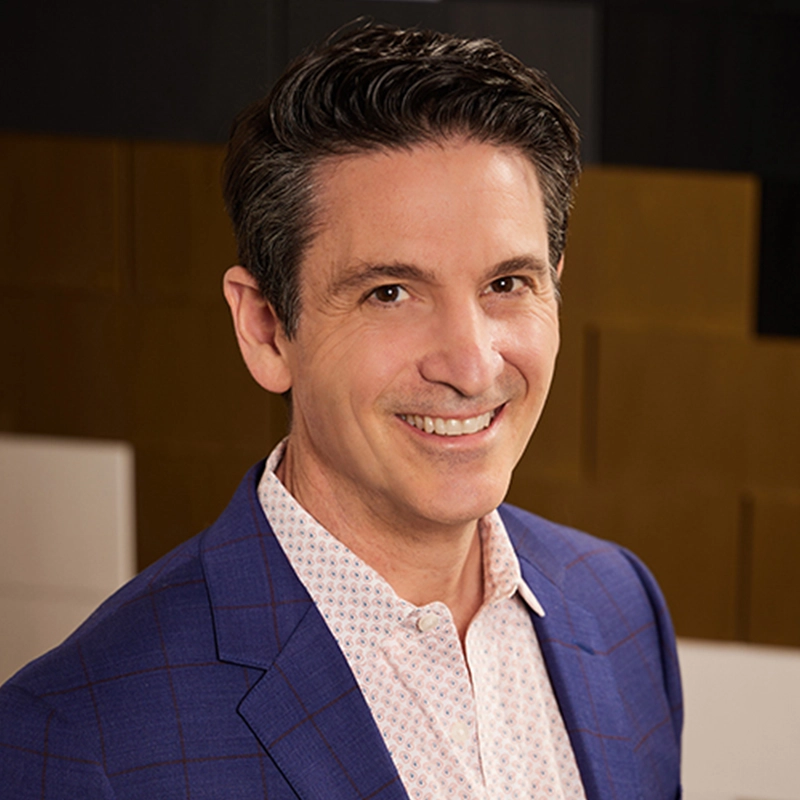 A man with short dark hair wearing a blue blazer over a light patterned shirt smiles at the camera. The well-lit, professional setting with brown and black geometric shapes in the background suggests an event featuring Lisette Glodowski.
