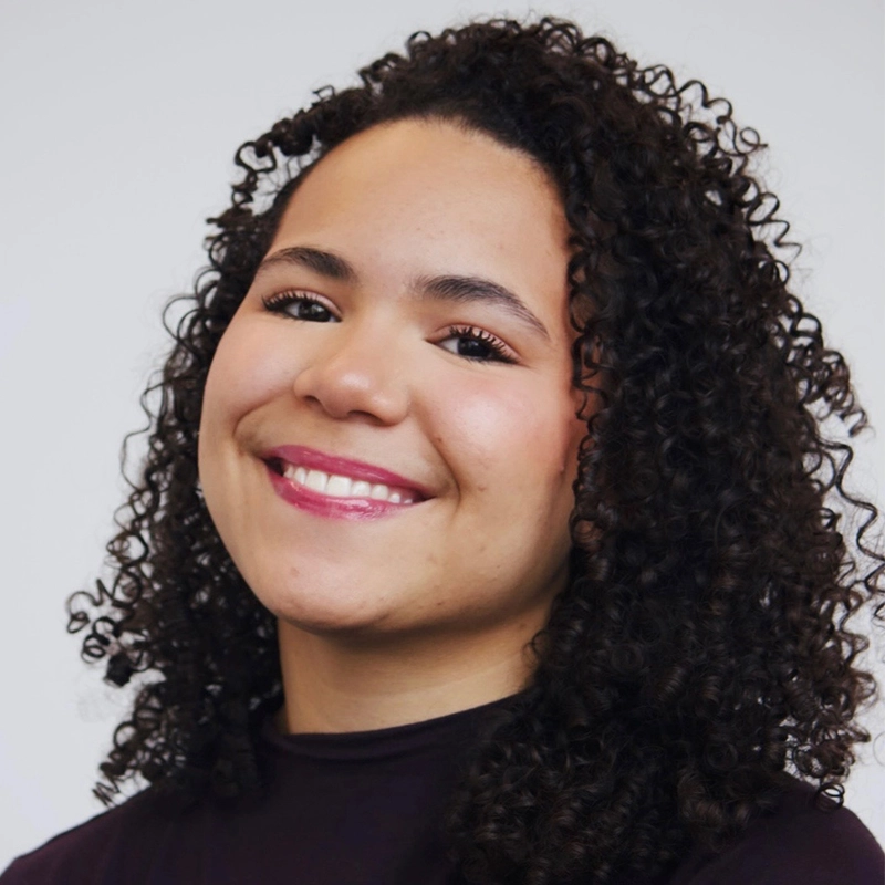 A woman with medium skin tone and curly dark hair smiles gently. She wears light makeup, pink lipstick, and a dark top. The plain, light-colored background creates a professional yet friendly atmosphere for this portrait image.