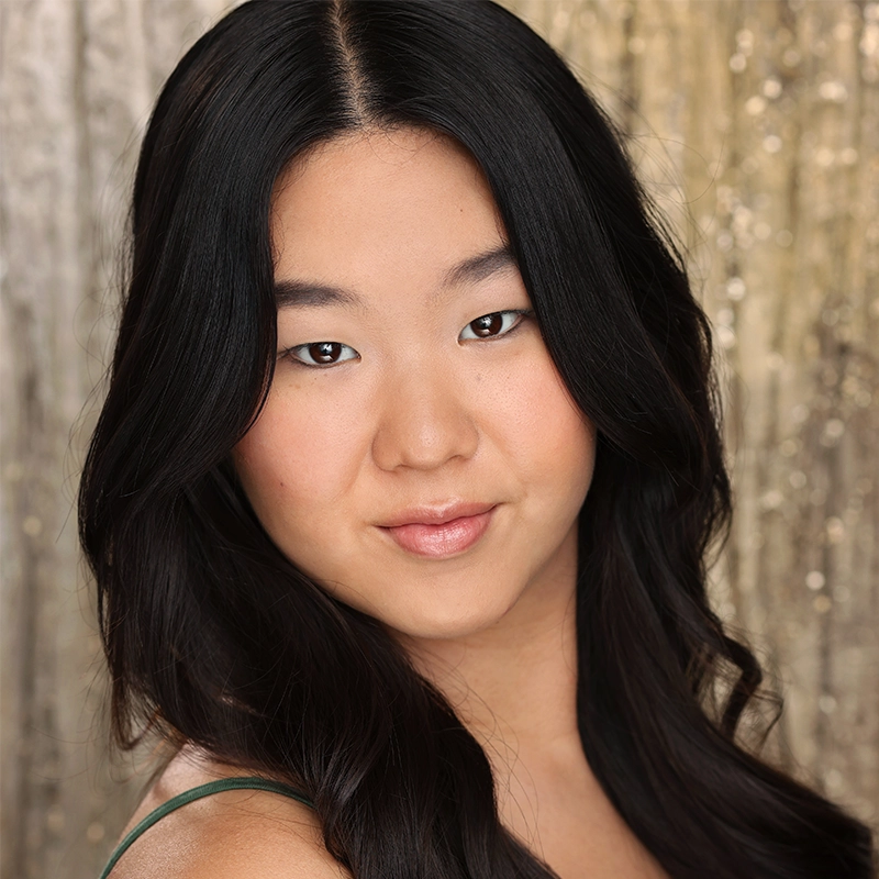 A young woman with long, wavy black hair and smooth skin looks confidently at the camera. She wears natural makeup and a green top. The shimmering background highlights her poise, reminiscent of Noah Ong Bamola’s distinctive style.