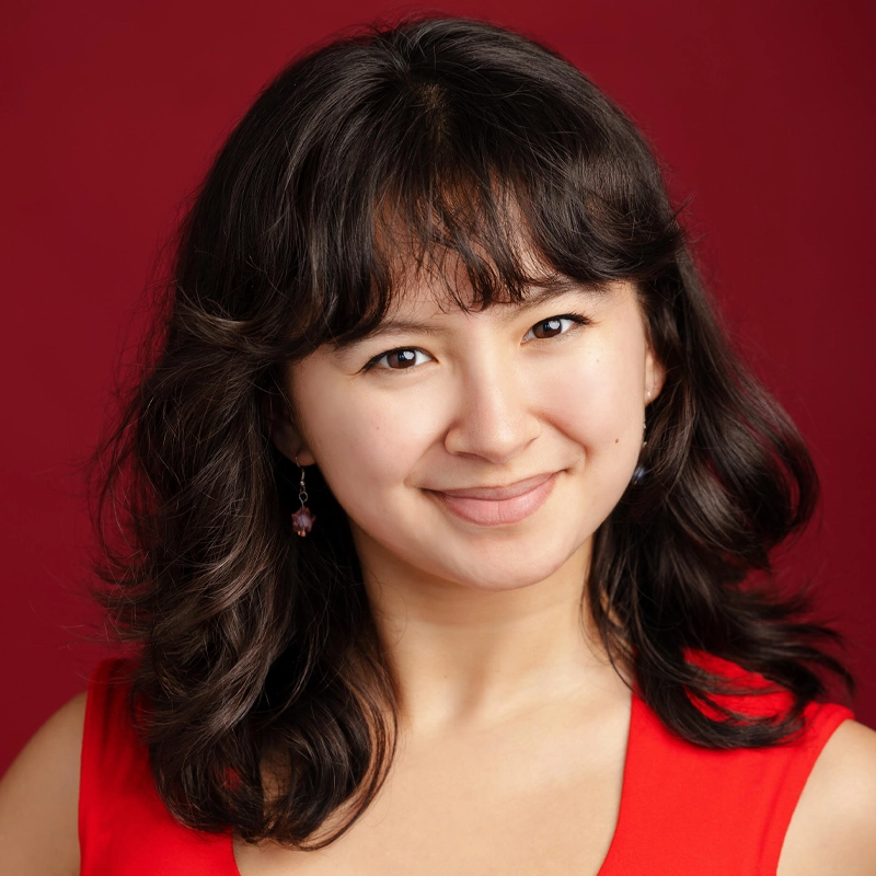 A woman with wavy, shoulder-length dark hair smiles softly. She wears a sleeveless red top and a single dangling earring. The background is solid red, complementing her outfit. Her expression is warm and approachable.