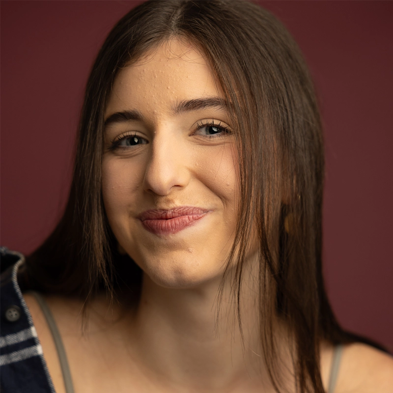 A young woman with long, straight brown hair smiles gently at the camera. She has light skin, blue eyes, and wears a gray top with a hint of a blue plaid shirt. The background is a deep burgundy shade.