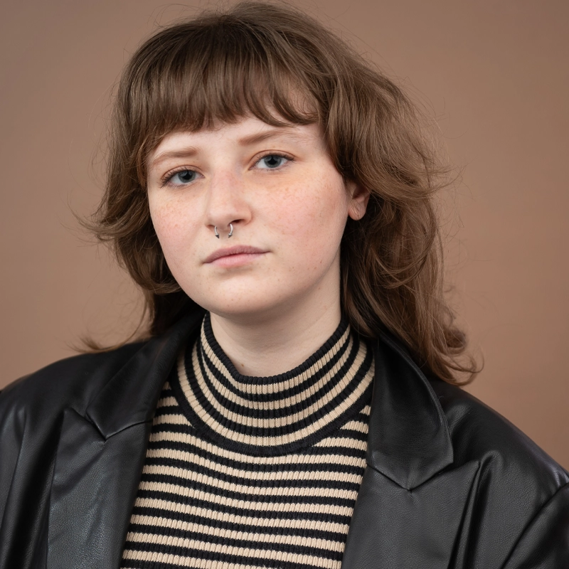 A young person with light skin, wavy brown hair, and a septum piercing looks at the camera. They wear a black and beige striped sweater under a black leather jacket. The background is solid brown. Their expression is calm and neutral, capturing Ciara Huckeby’s distinctive style.