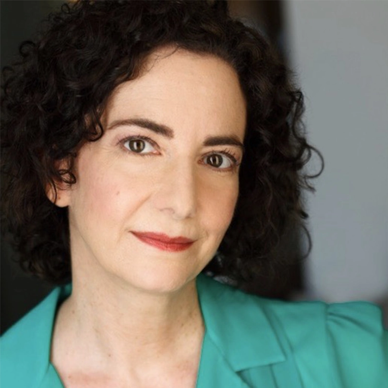 Lisette Glodowski, a woman with short, dark curly hair and fair skin, looks at the camera with a faint smile. She wears a teal blazer and subtle makeup with red lipstick. The background is softly blurred in neutral tones.