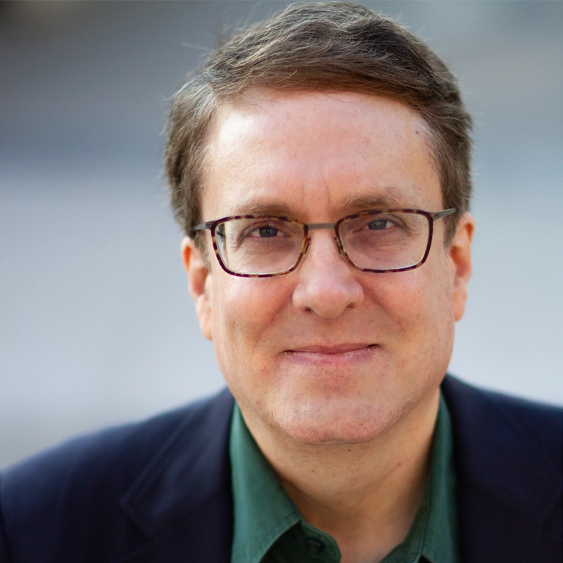 A middle-aged man with short brown hair and glasses smiles gently at the camera. Wearing a dark blazer over a green collared shirt, he stands in sharp focus, the background softly blurred—possibly at an event featuring Lisette Glodowski.