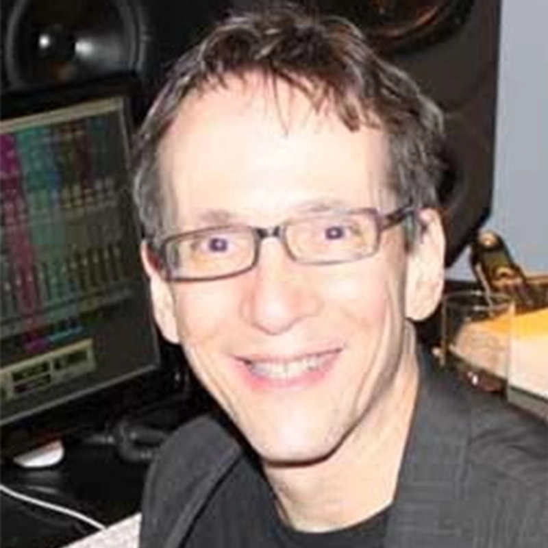 A smiling man with short brown hair and glasses sits in front of audio equipment and a computer screen displaying colorful sound waveforms in a recording studio, working on a project with Lisette Glodowski. He is wearing a dark jacket and black shirt.