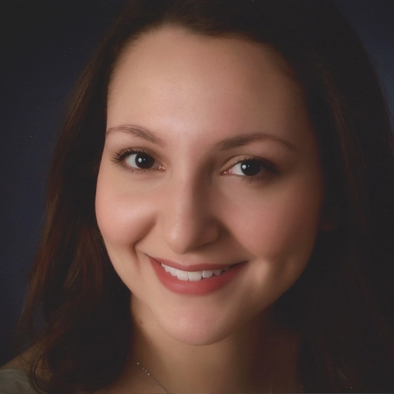 Lisette Glodowski, with long brown hair and brown eyes, smiles at the camera. She has clear skin, visible dimples, and wears subtle makeup. The dark blue, softly lit background highlights her face.