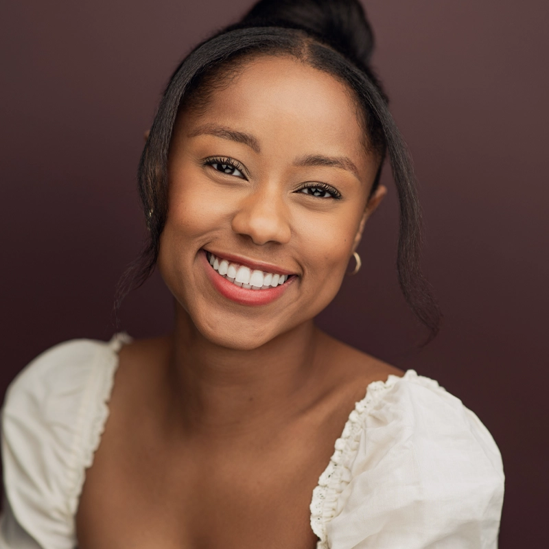 Kaléa Leverette, a young woman with medium brown skin and dark hair in a bun, smiles warmly at the camera. She wears small hoop earrings and a white, square-neck blouse with puffed sleeves, set against a solid maroon background.