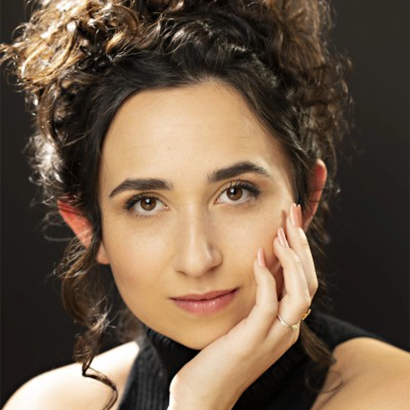 Lisette Glodowski, with her curly brown hair styled up, poses with her chin resting on her hand. She has fair skin, brown eyes, and wears a sleeveless black top. Her calm, confident expression stands out against the dark, softly blurred background.