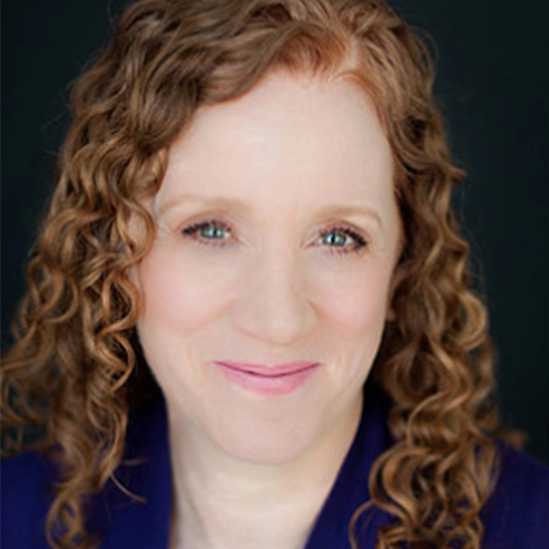 Lisette Glodowski, a woman with fair skin and blue eyes, smiles gently. She has long, light brown curly hair and is wearing a dark blue top. The dark background makes her face and hair stand out clearly in the image.