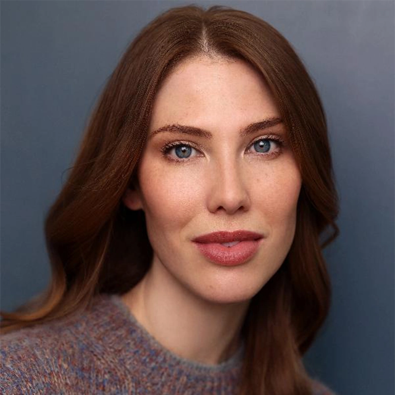 Tess Marshall, a woman with fair skin, long brown hair, and blue eyes, looks at the camera wearing a soft, grayish sweater. She has minimal makeup and a gentle expression, with a blurred blue-gray background behind her.