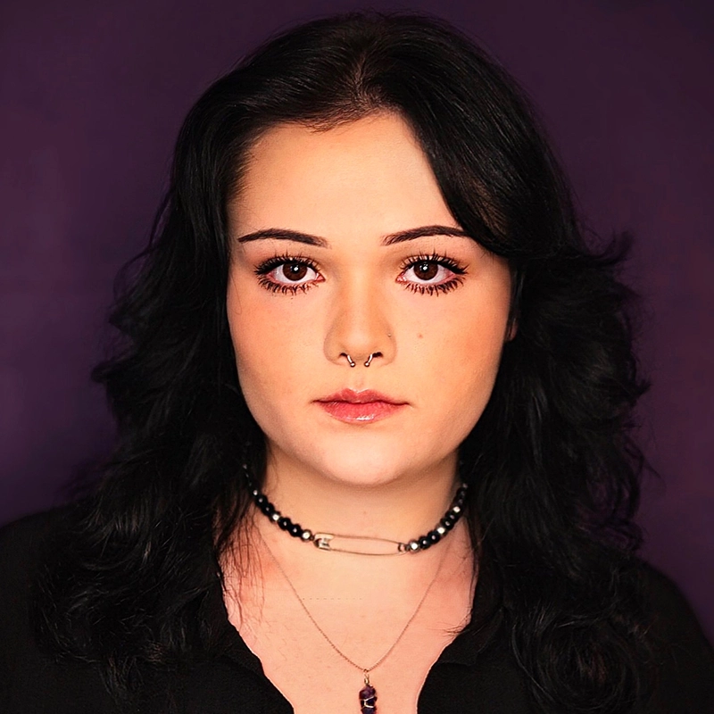 A young woman with wavy black hair and fair skin poses against a solid purple background. She wears subtle makeup, a septum ring, layered necklaces, and a black top, looking directly at the camera with a neutral expression.
