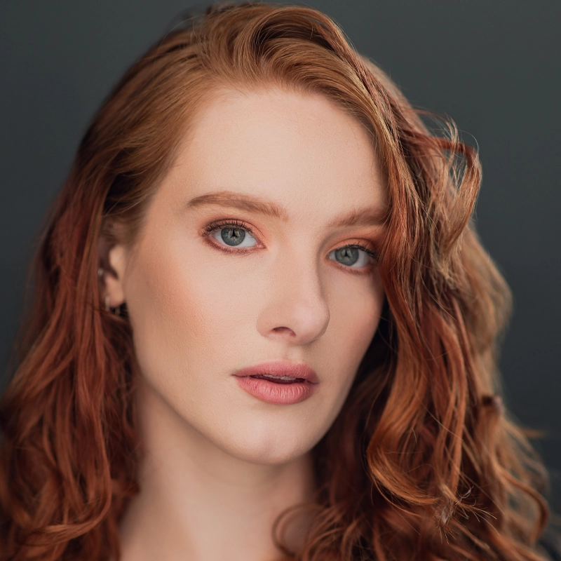 Close-up portrait of Jessica Vanek, a woman with fair skin and long, wavy red hair. She has blue eyes, soft natural makeup with peachy tones, and a neutral expression against a plain, dark gray background that keeps the focus on her face.
