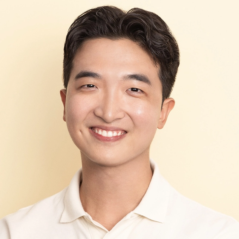 A young man with short, dark hair smiles at the camera. He is wearing a cream-colored collared shirt and stands against a soft beige background. His expression is friendly and relaxed, with even lighting across his face.