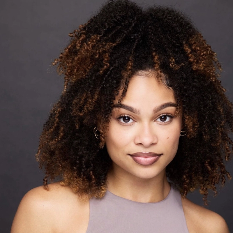 Dasia Alonna Amos, a woman with medium brown skin and voluminous curly hair with blonde highlights, looks directly at the camera. She wears a sleeveless, high-necked gray top and small hoop earrings against a dark gray background.